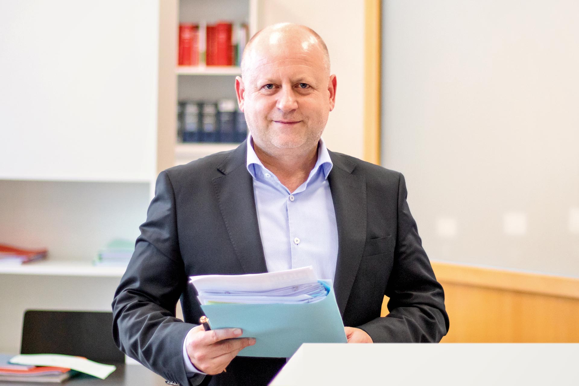 Ein Mann in einem B&uuml;ro h&auml;lt Akten und l&auml;chelt in die Kamera, B&uuml;cher sind im Hintergrund auf einem Regal.